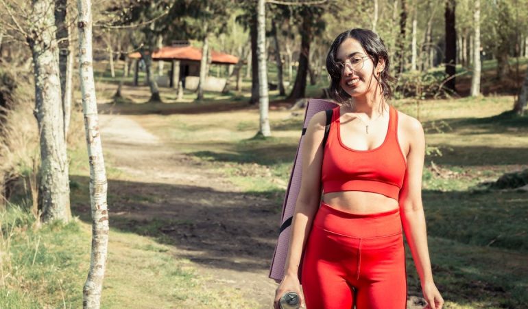 Smiling woman in red sport clothing holding a yoga mat outdoors.